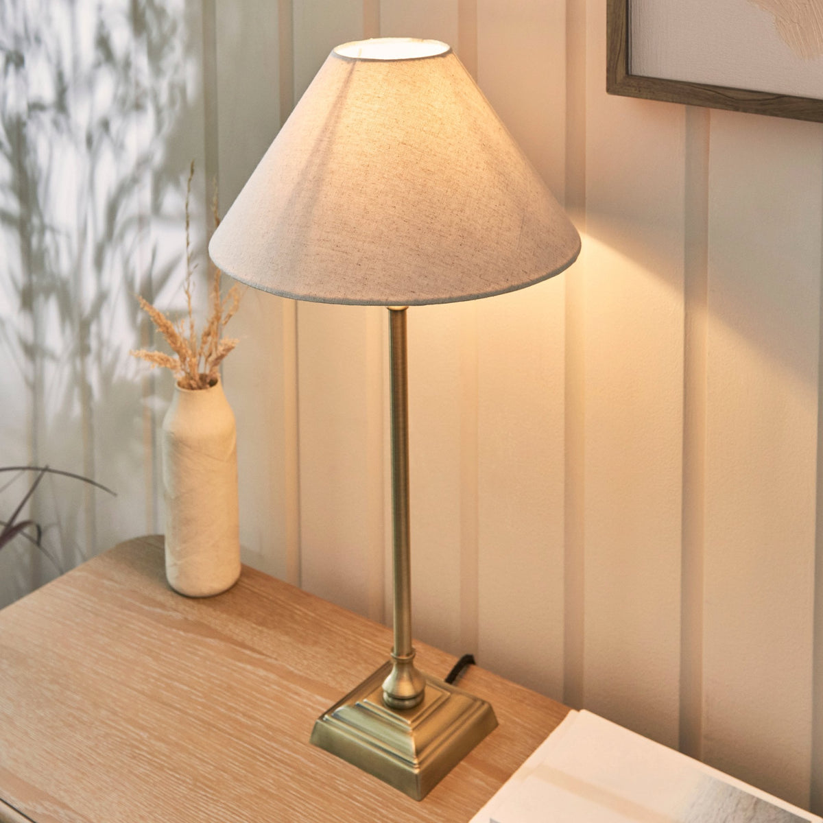 A brass table lamp with a linen shade on a wooden desk, next to a vase with dried grass.