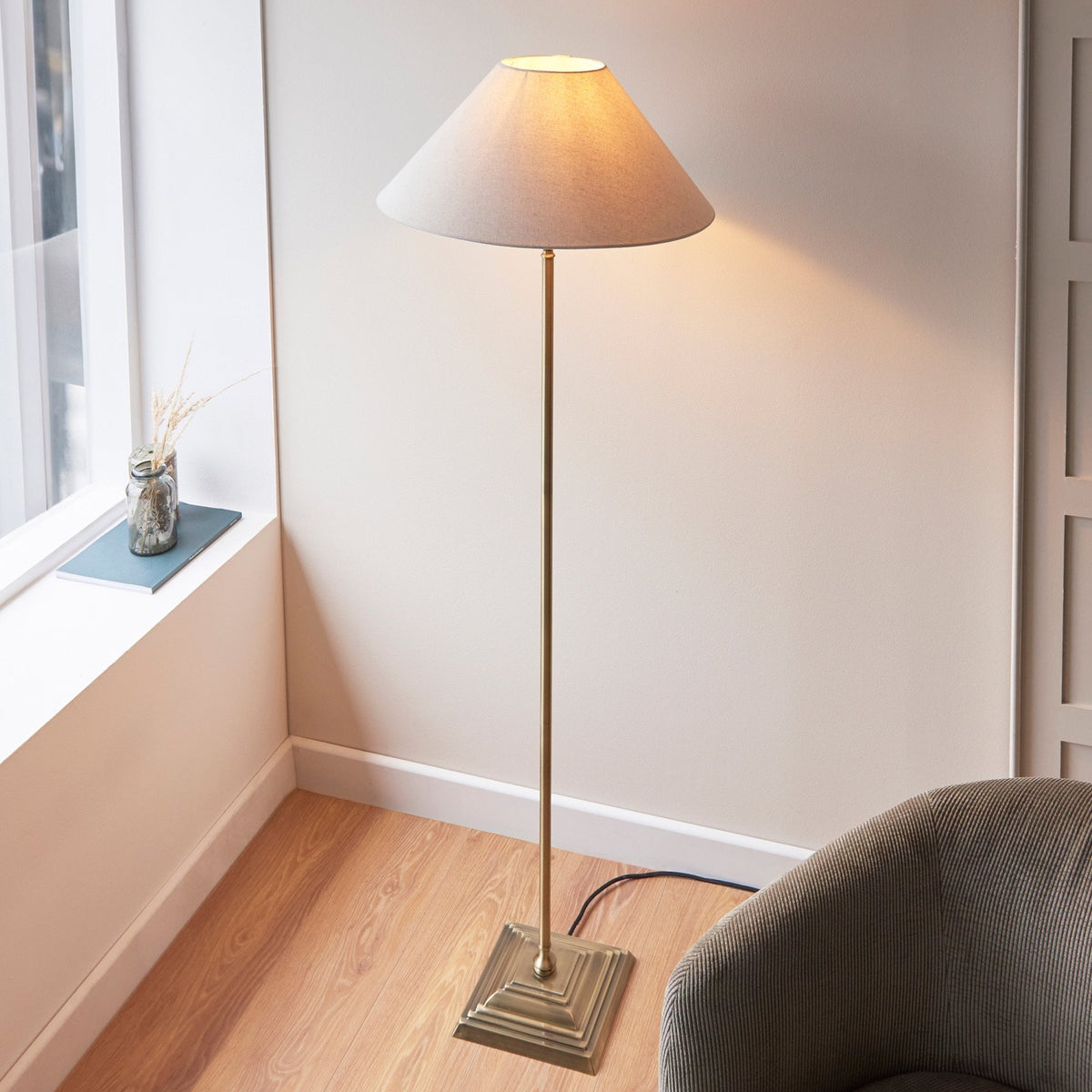 A brass floor lamp with a cream shade stands in a bright room next to a window and a chair.