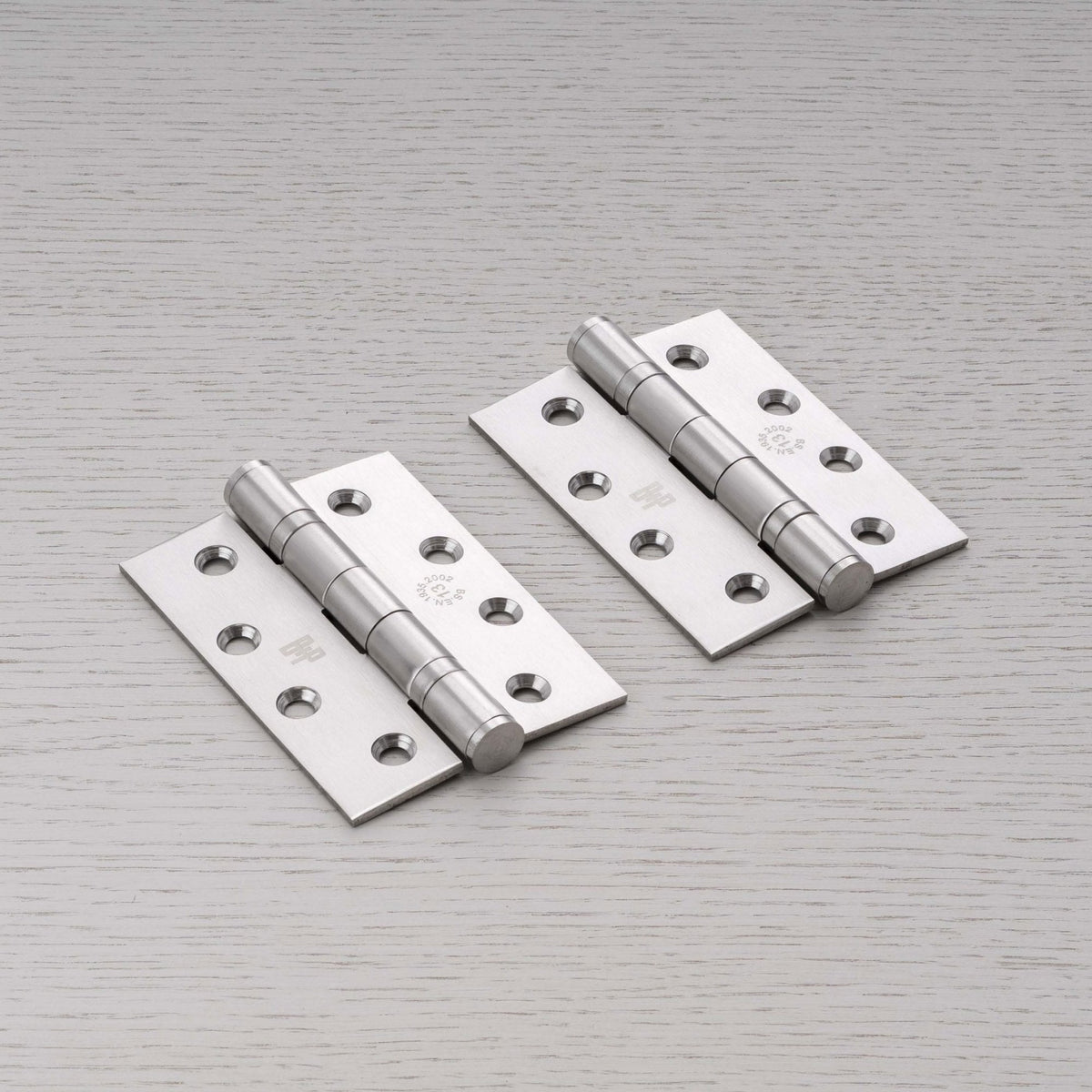 Pair of satin chrome door hinges with fixing holes on a light wood surface.