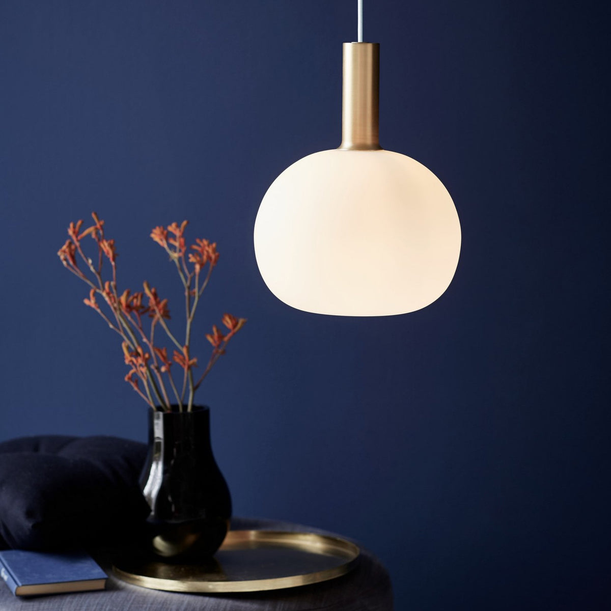 A large white pendant light with a brass neck, above a side table with a vase and book.