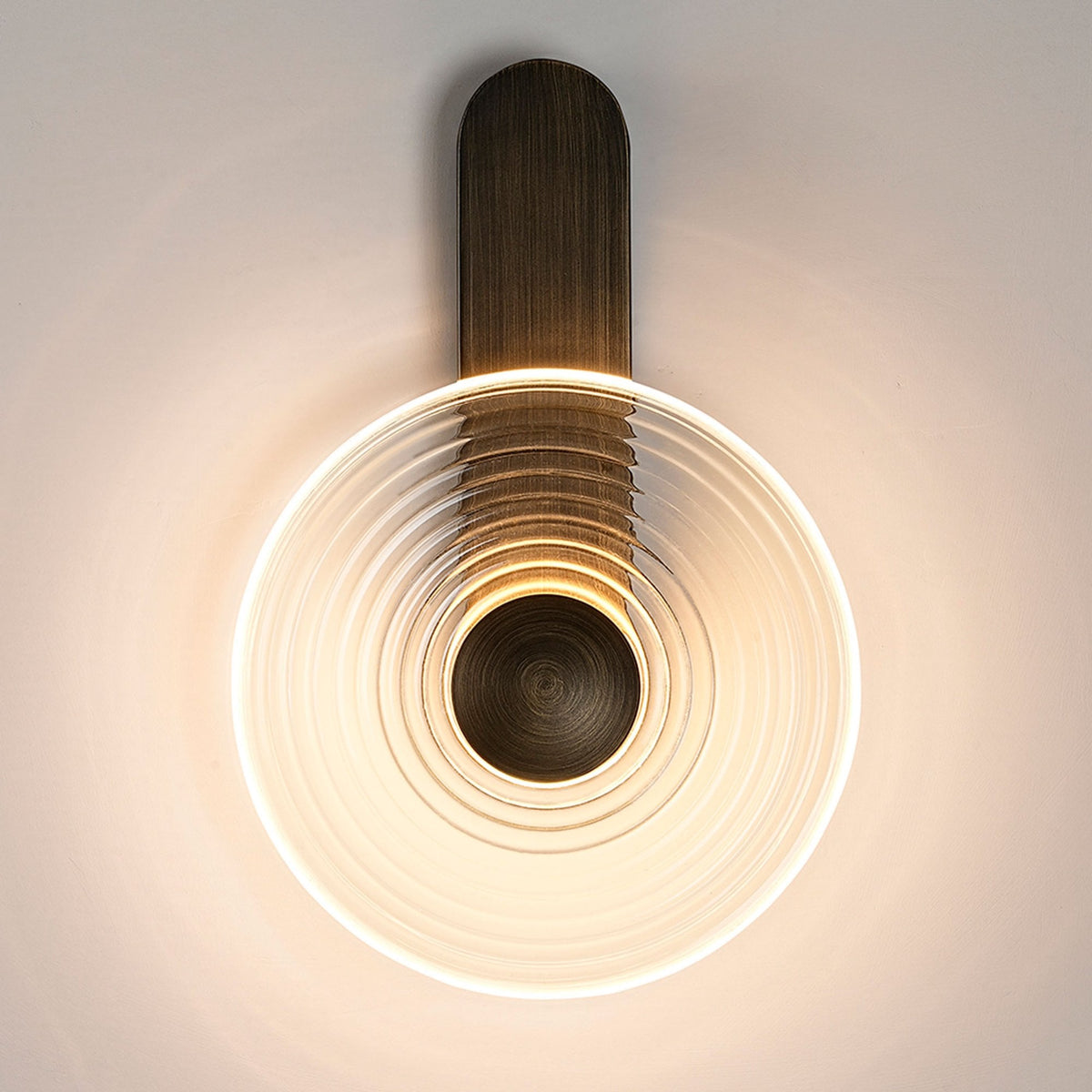 A modern wall lamp with a round, clear, ribbed glass shade and a dark metal base, lit.