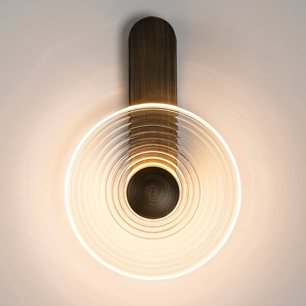 A modern wall lamp with a round, clear, ribbed glass shade and a dark metal base, lit.