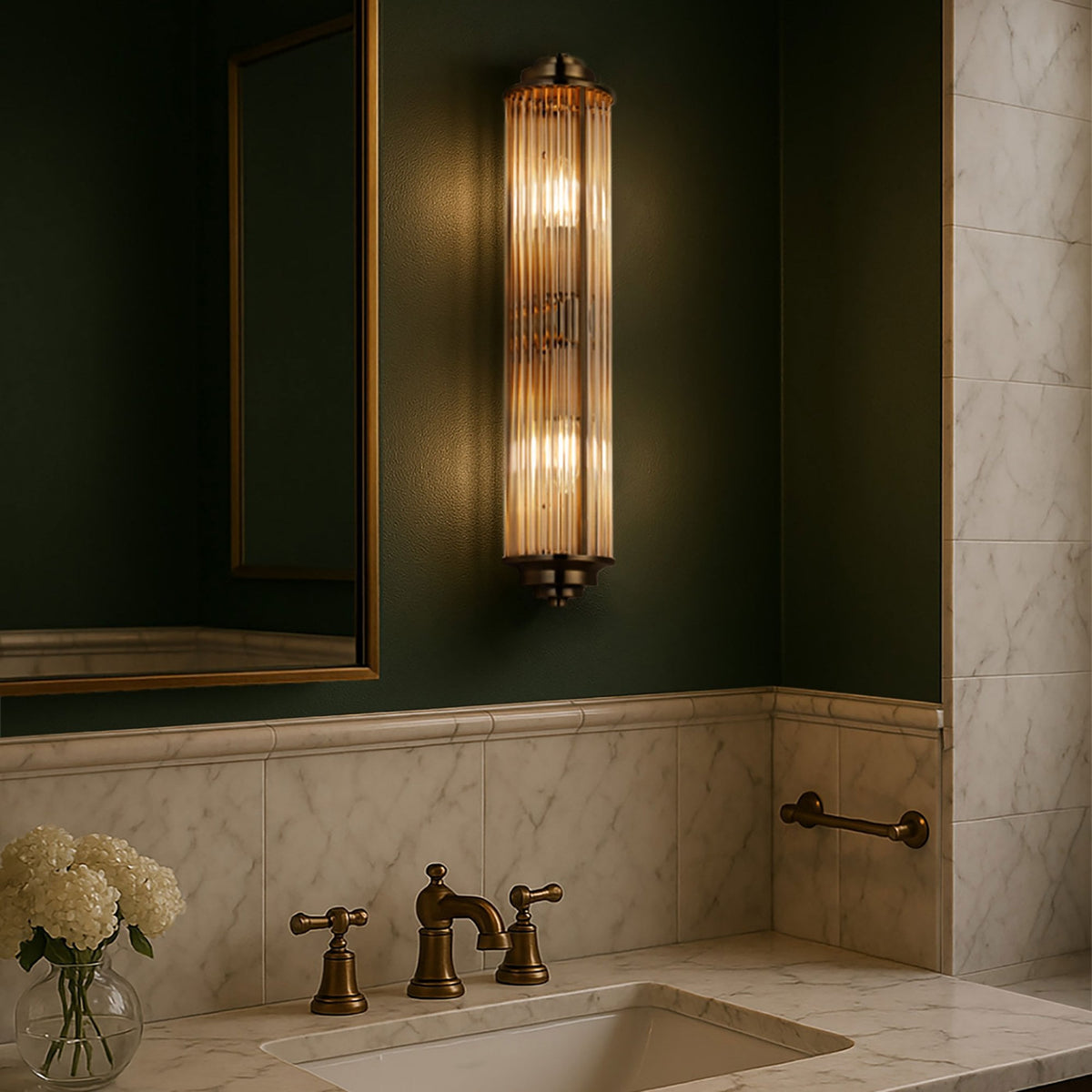 A brushed brass reeded glass wall light illuminates a dark green bathroom wall above a marble sink.