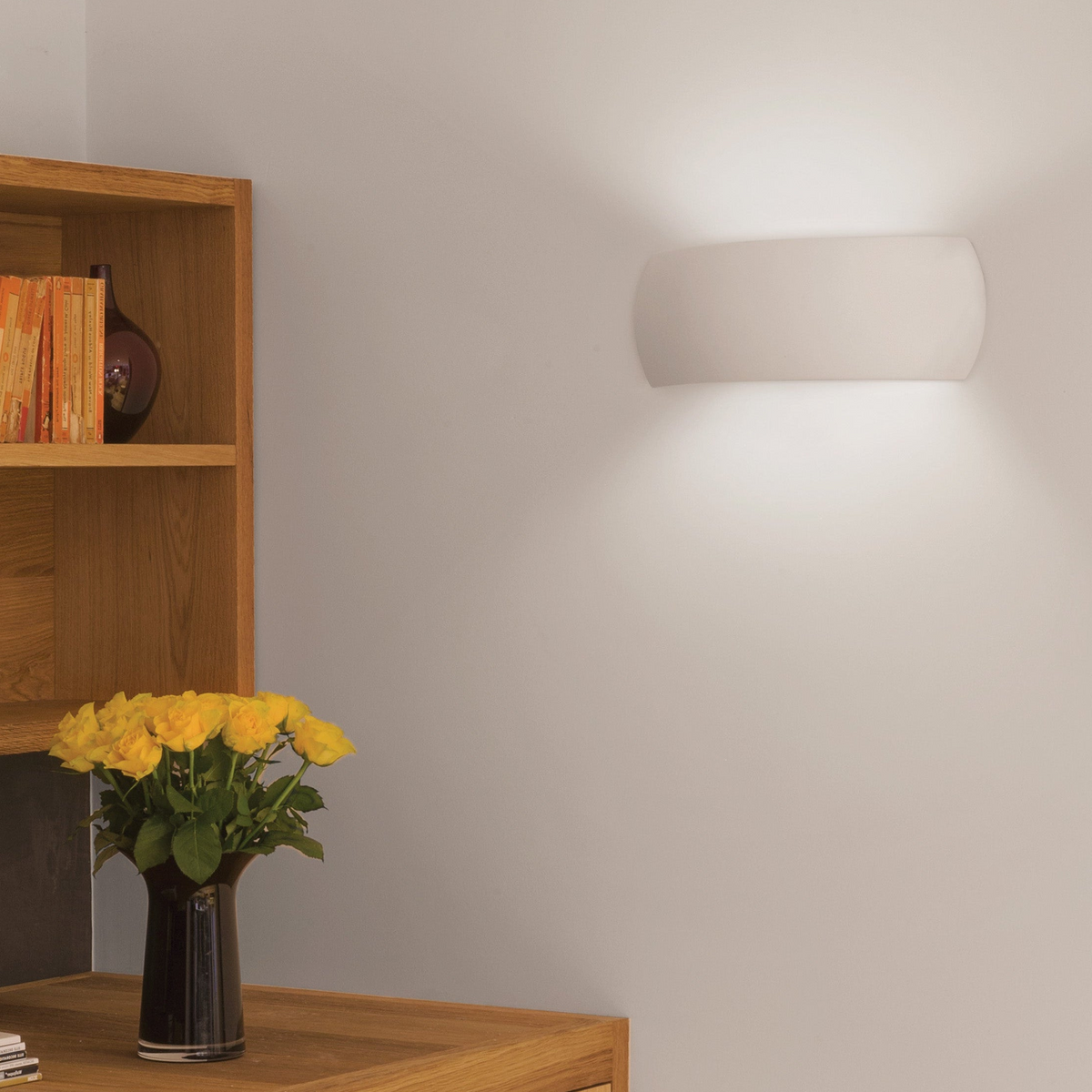 White wall light illuminating a wall above a wooden shelf with books and yellow roses in a vase