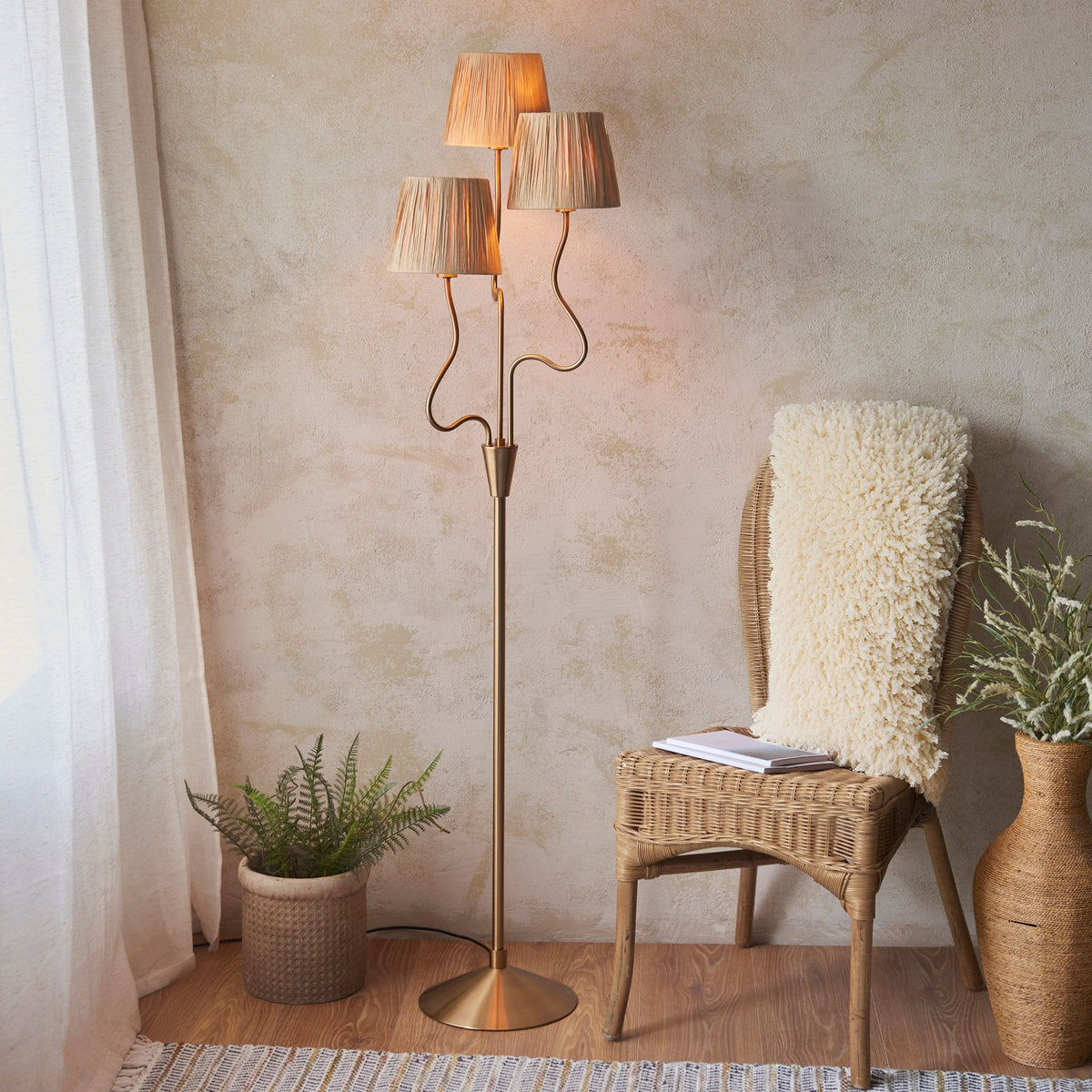 A tall, brass floor lamp with three pleated raffia shades, illuminating a cozy living space with a rattan chair and plants.