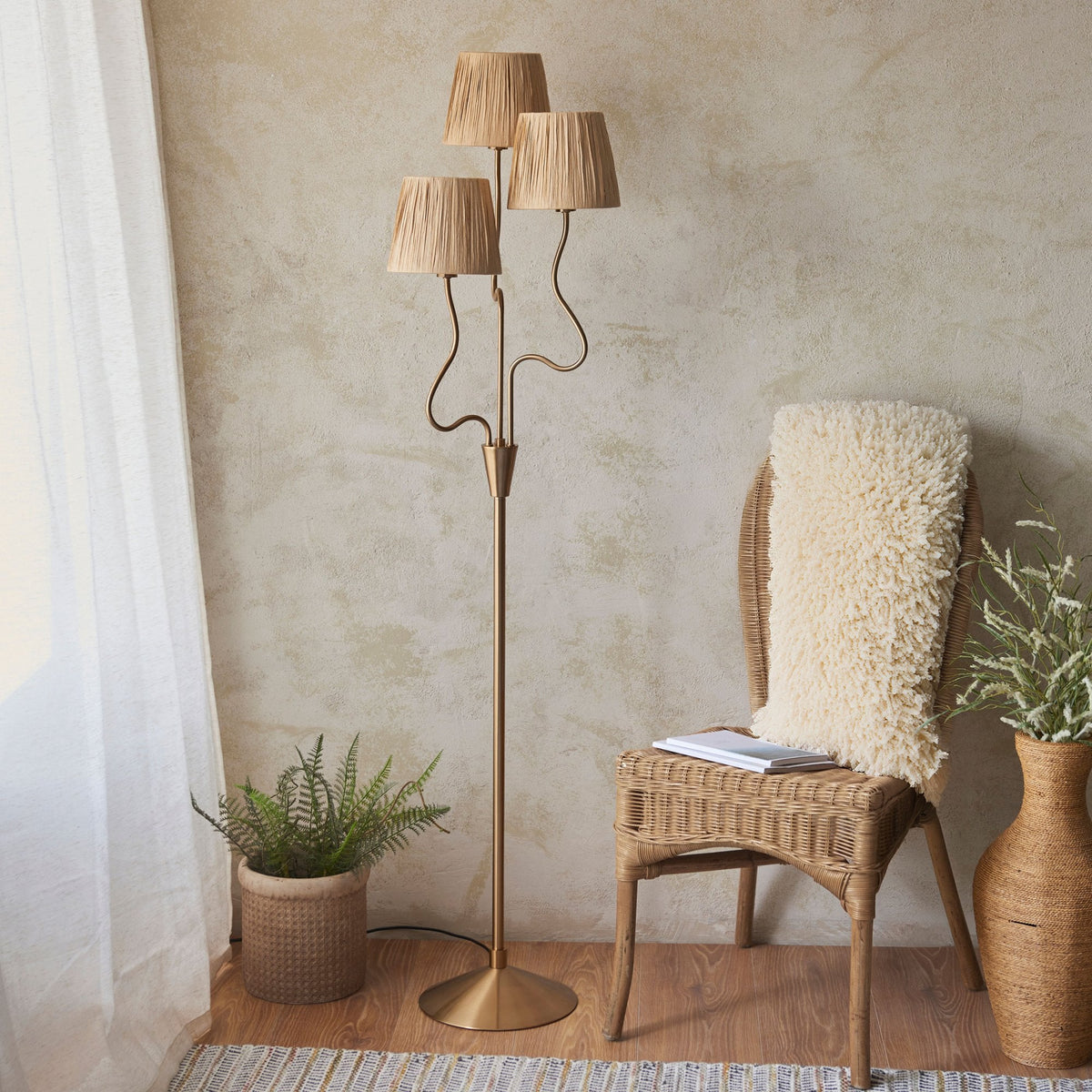 A tall, brass floor lamp with three pleated raffia shades, illuminating a cozy living space with a rattan chair and plants.
