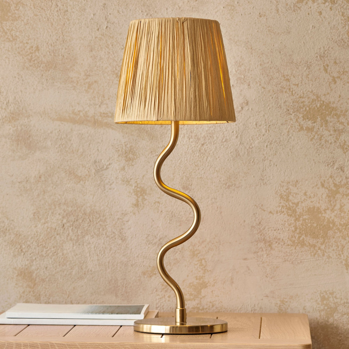 A gold table lamp with a wavy stem and a pleated beige shade sits on a light wooden table.