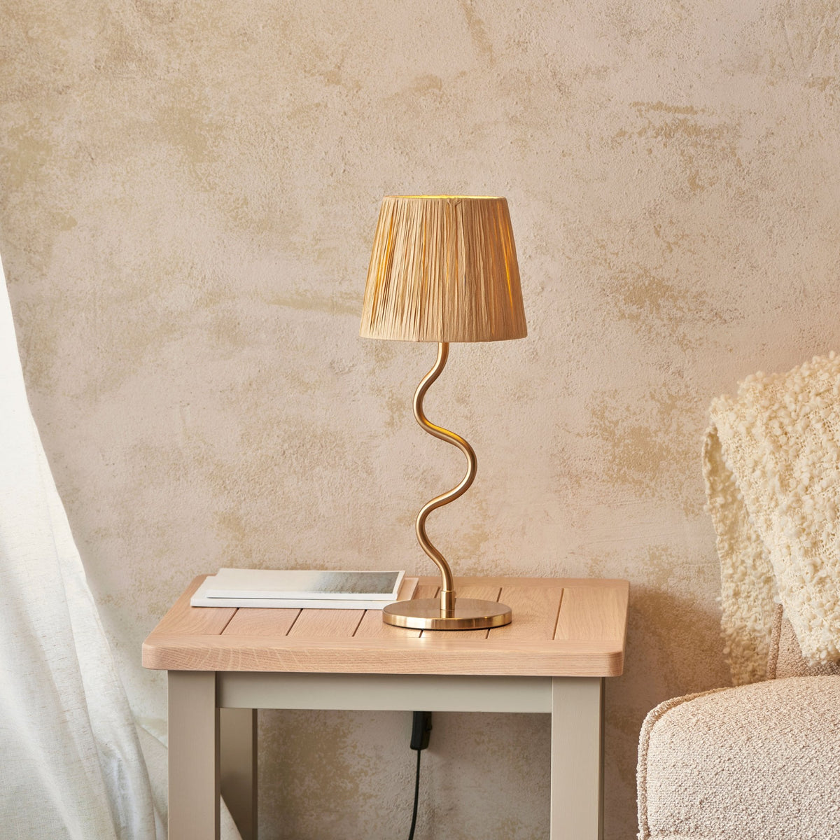 A gold table lamp with a wavy stem and a pleated beige shade on a light wooden nightstand next to a bed.
