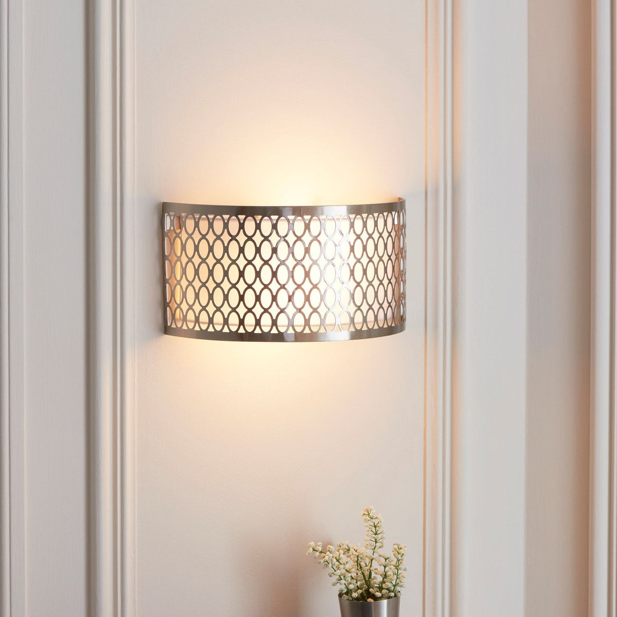 A brushed chrome wall light with an intricate oval pattern, casting a warm glow on a white wall.
