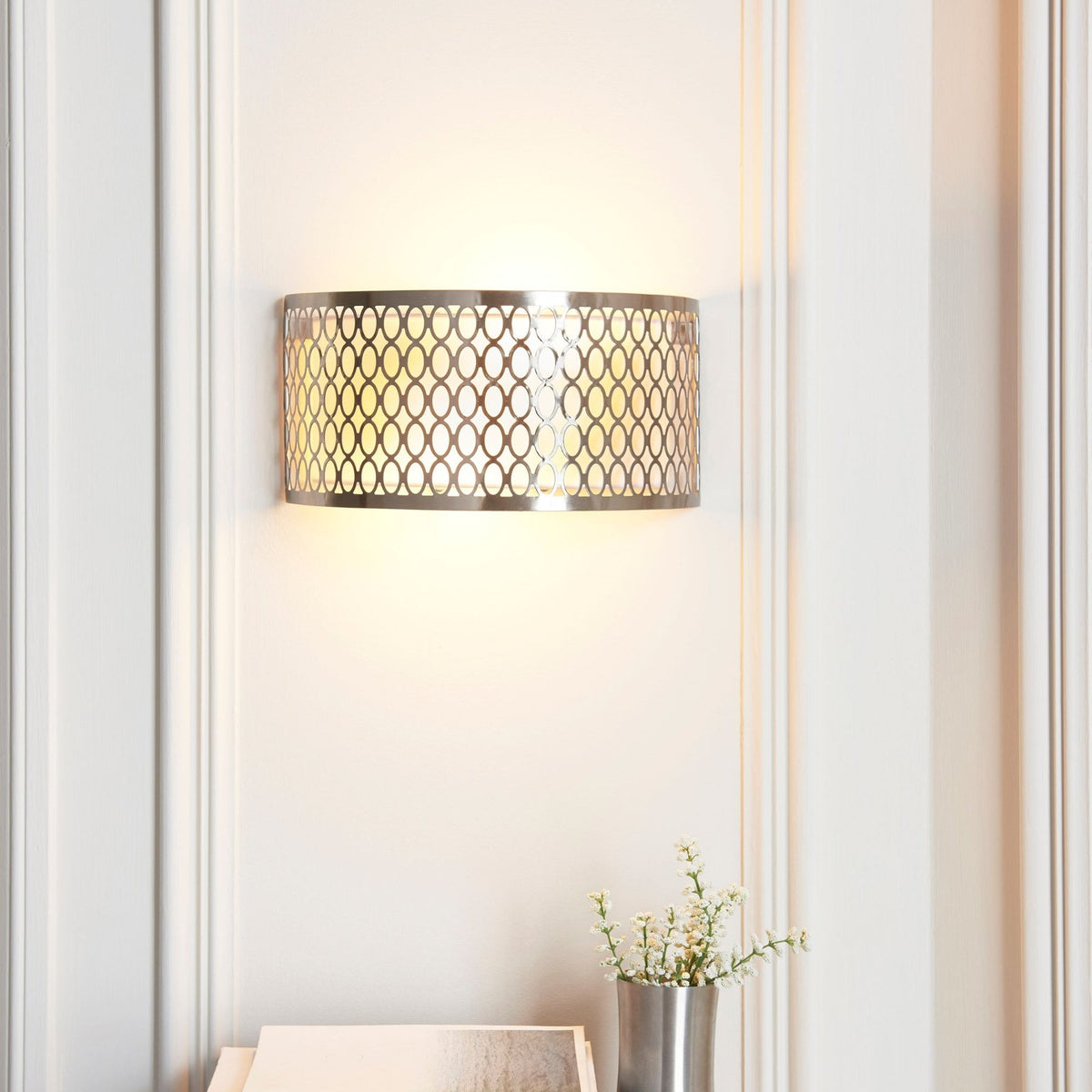 A brushed chrome wall light with an intricate oval pattern, casting a warm glow on a white wall above a plant.