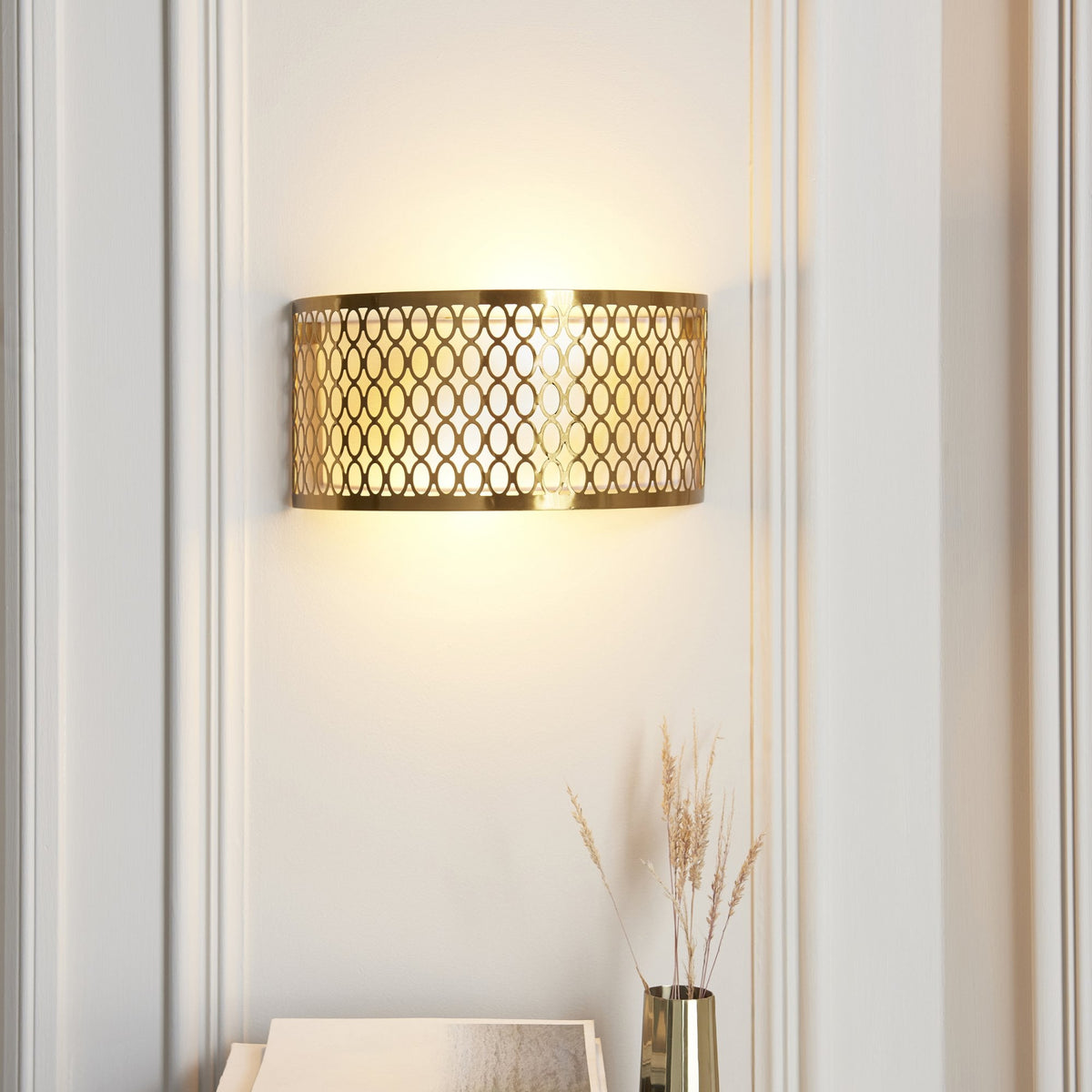 Gold wall light with an intricate oval pattern, casting a warm glow on a white paneled wall.