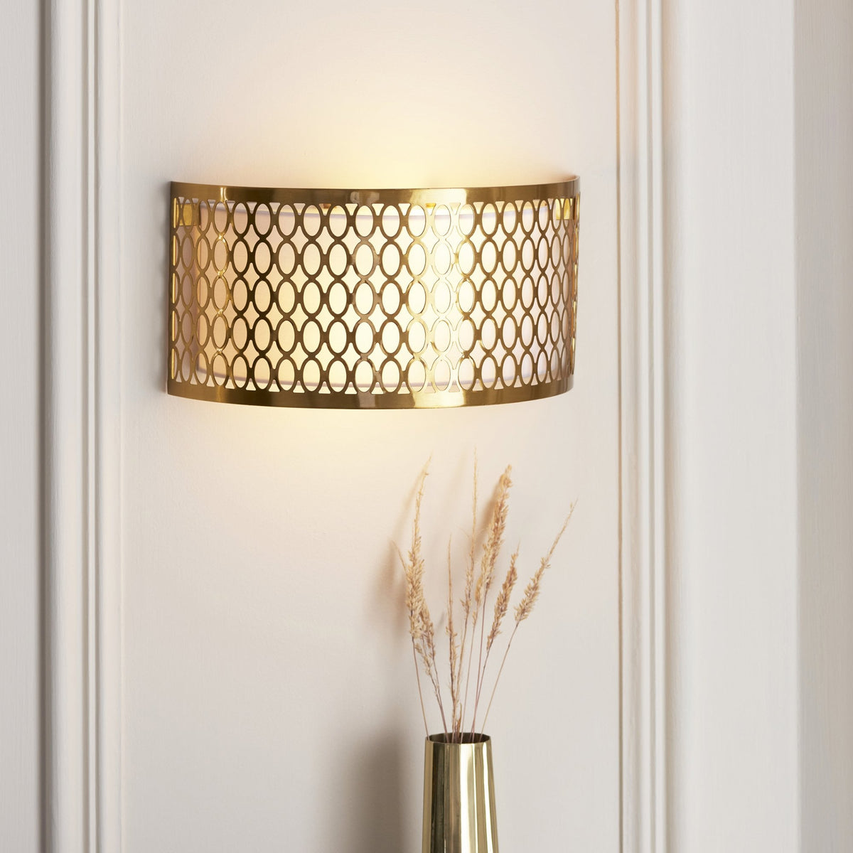 Gold wall light with an intricate oval pattern illuminating a white wall, with dried pampas grass in a gold vase.
