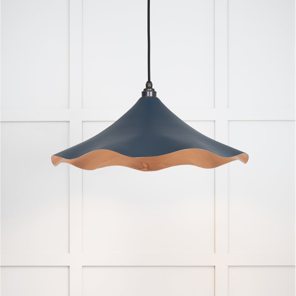 A dark blue and copper wavy-rimmed pendant light suspended against a white panelled wall.
