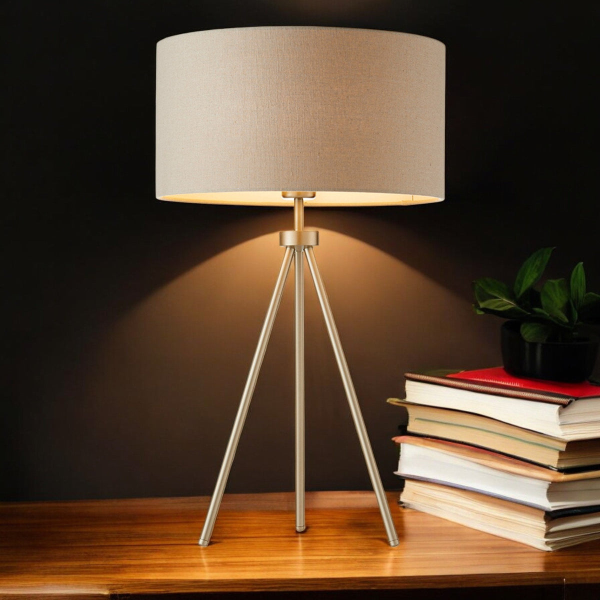 Tripod table lamp with a beige shade and gold base, illuminating a stack of books and a small plant.