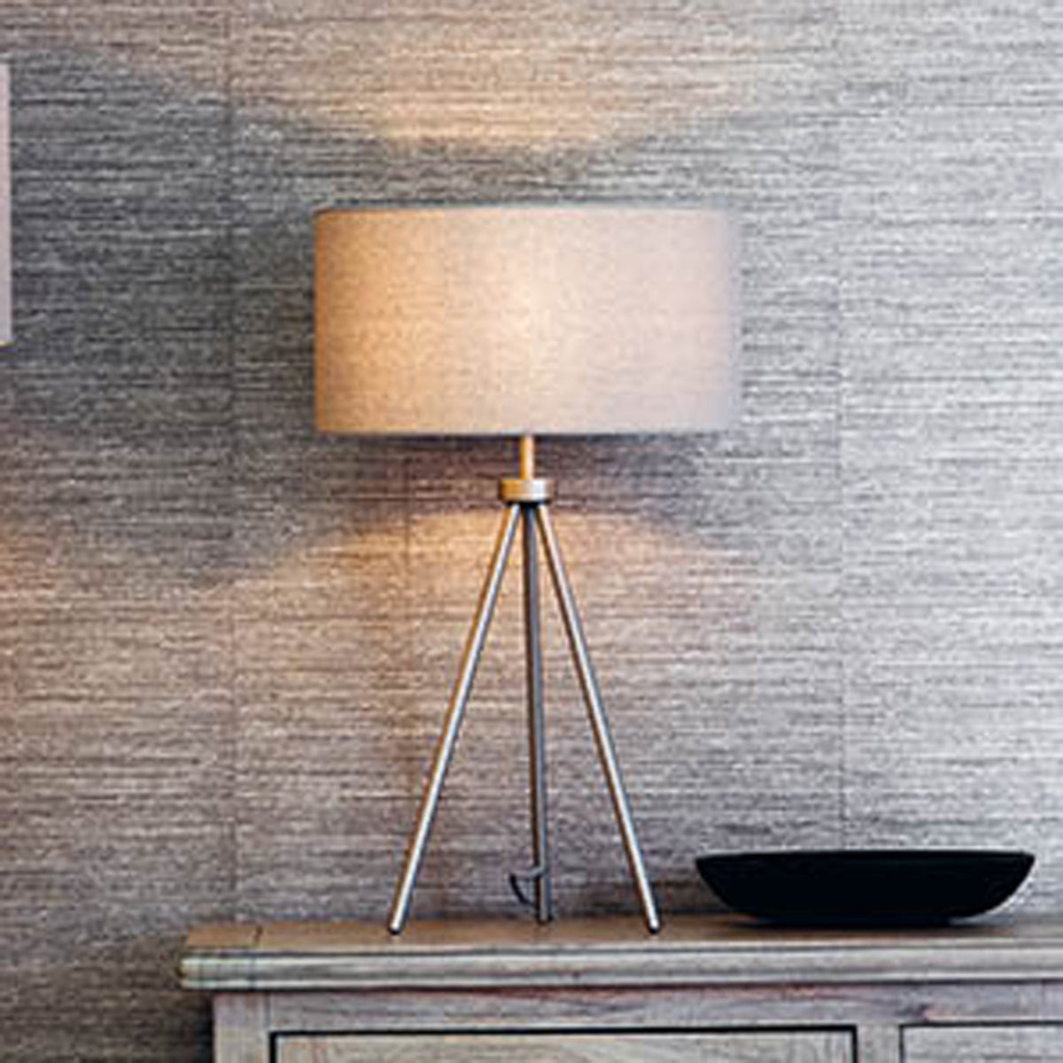 Tripod table lamp with a beige shade and gold base on a wooden surface next to a black bowl.