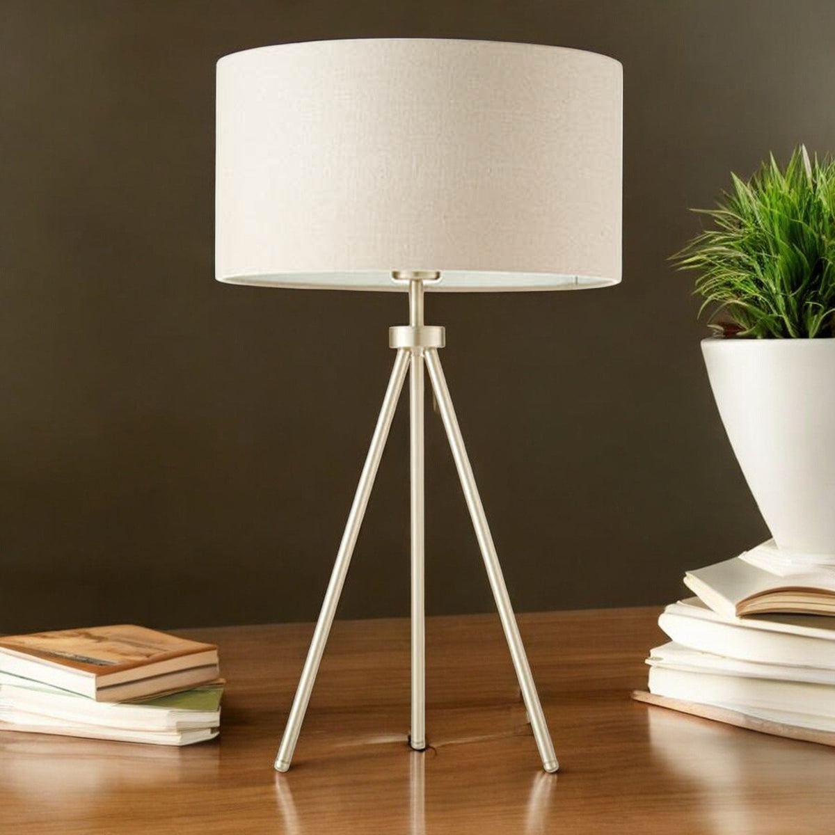 Tripod table lamp with a beige shade and gold base on a wooden desk with books and a plant.
