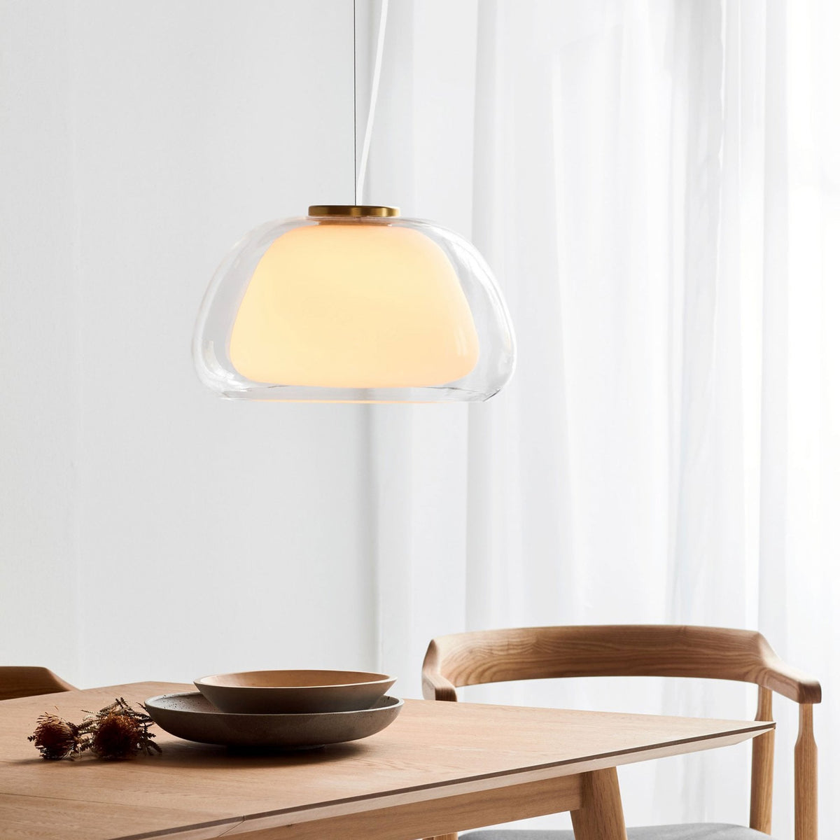 An illuminated pendant light hangs over a wooden table set with plates, in a light-filled room.