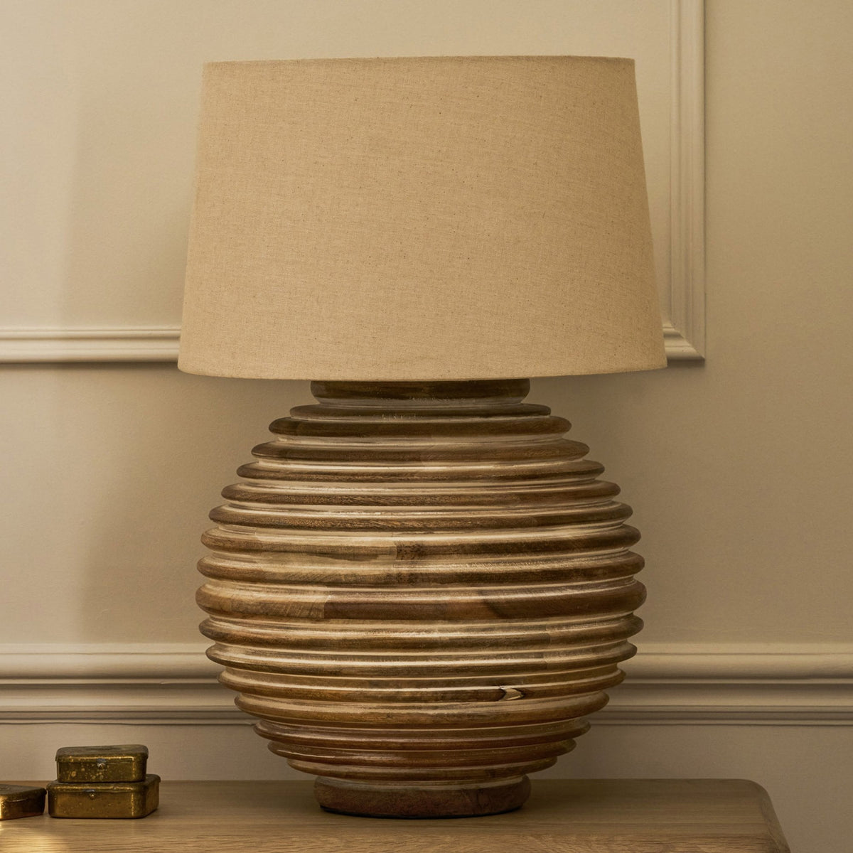 A wooden table lamp with a textured base and a light brown fabric shade, on a wooden surface.