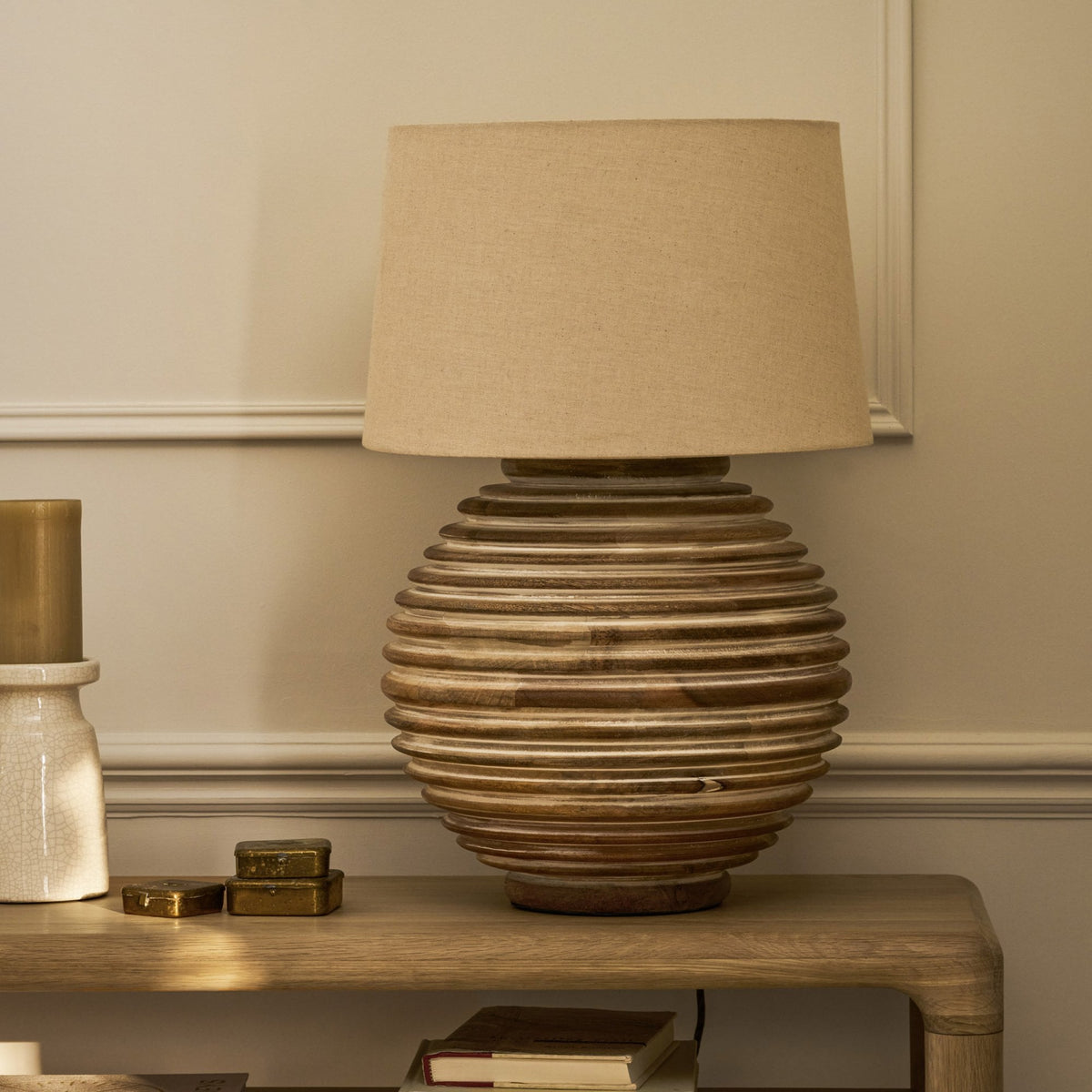 A wooden table lamp with a beige shade and a ribbed base, next to a gold candle and small brass boxes.