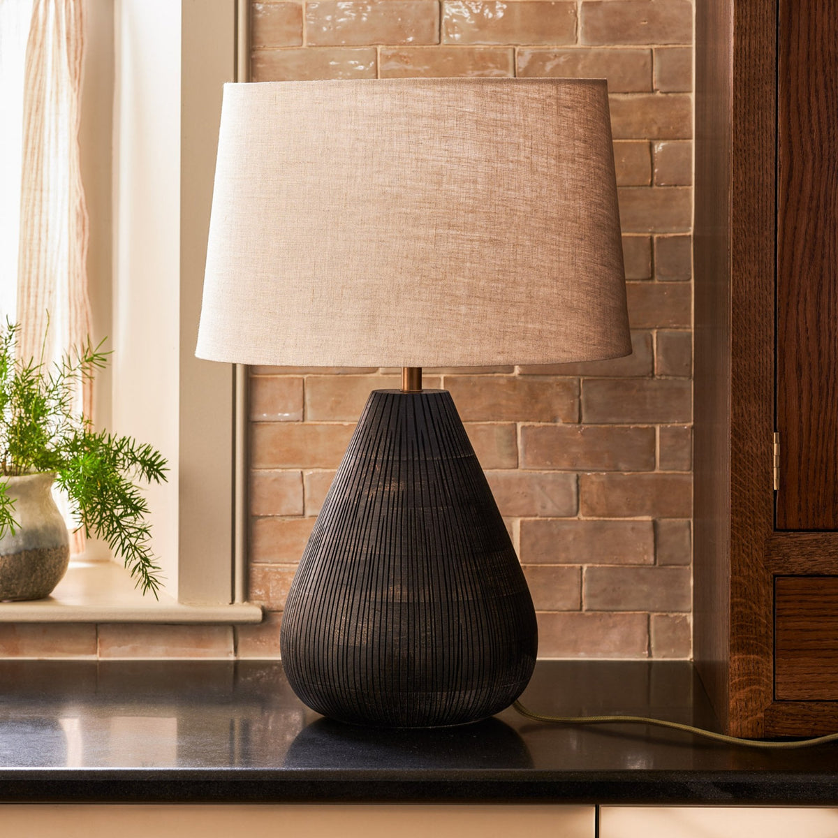 A black textured lamp with a light fabric shade sits on a dark countertop, next to a window and a plant.