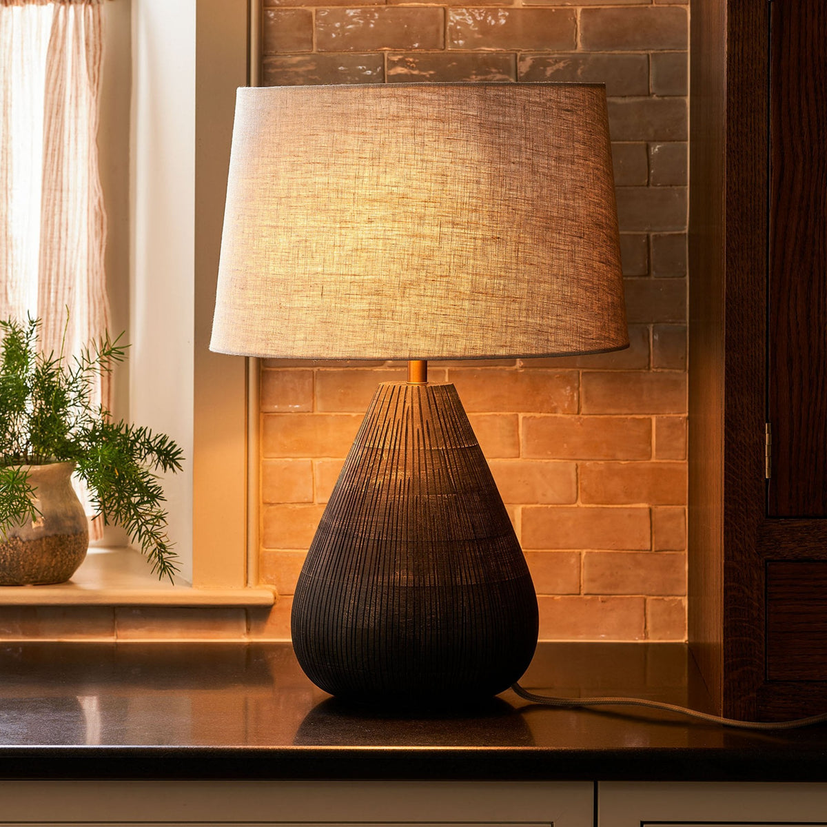 A black textured table lamp with a light linen shade sits on a dark countertop, illuminating a brick wall.