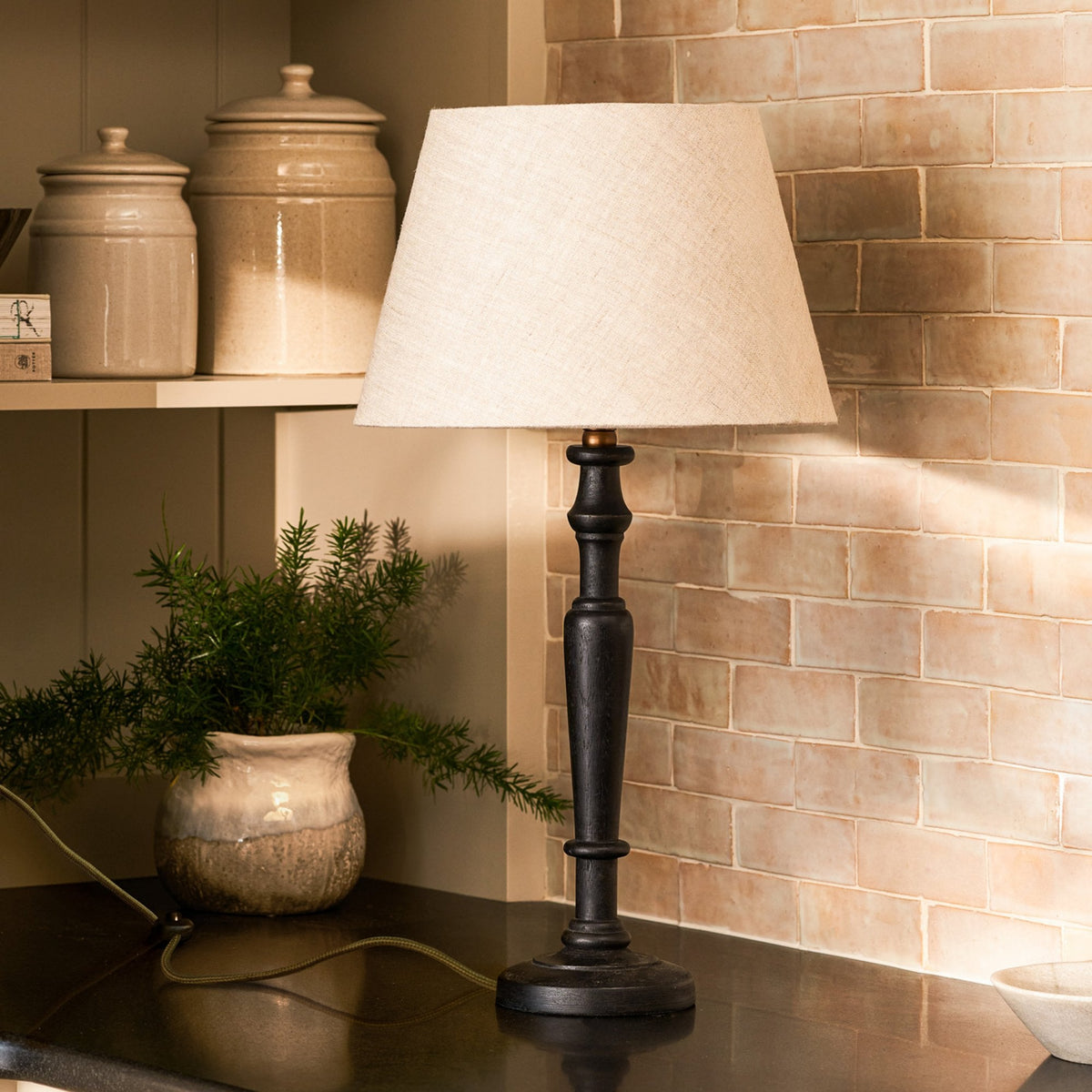 A black wooden table lamp with a cream linen shade on a dark countertop, next to a potted plant.