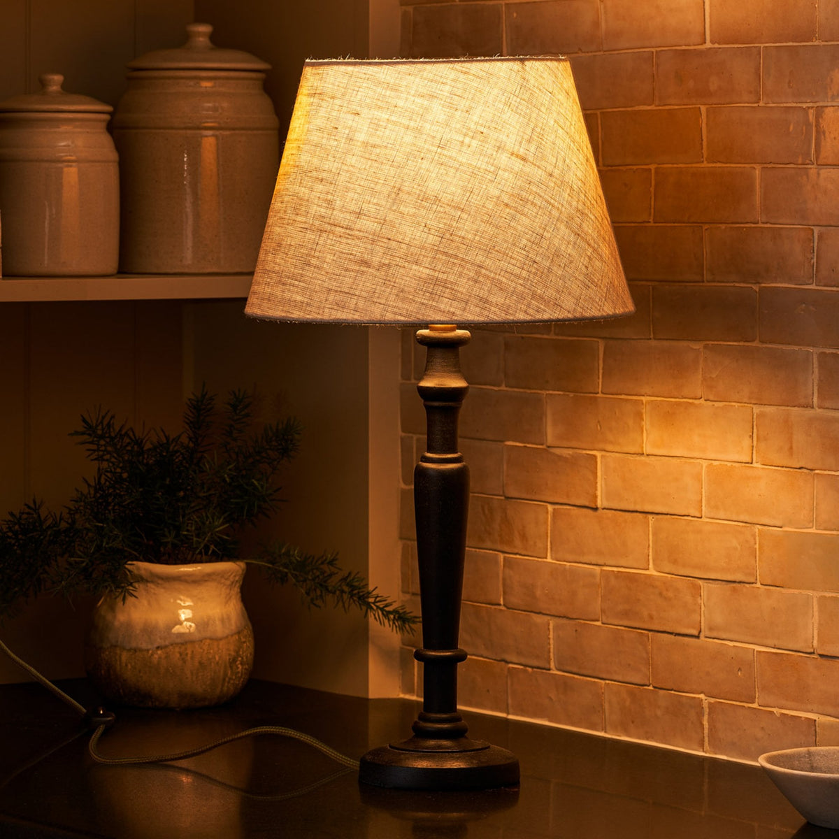 A rustic table lamp with a linen shade illuminates a kitchen counter with warm light.