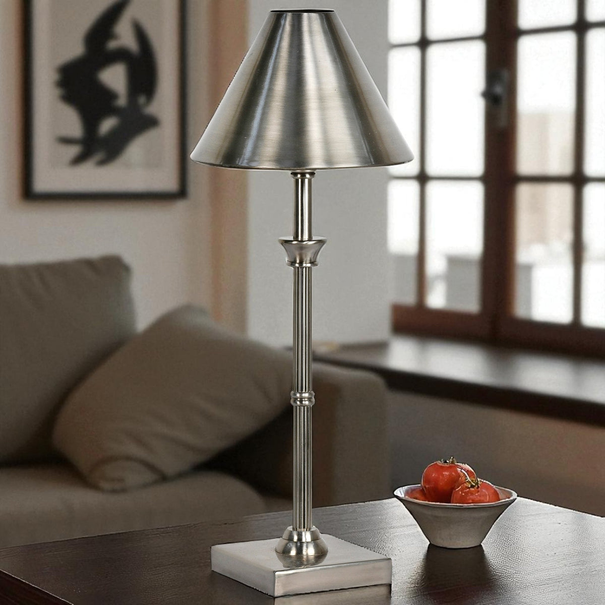 A brushed nickel table lamp with a conical shade sits on a dark wood table next to a bowl of pomegranates.