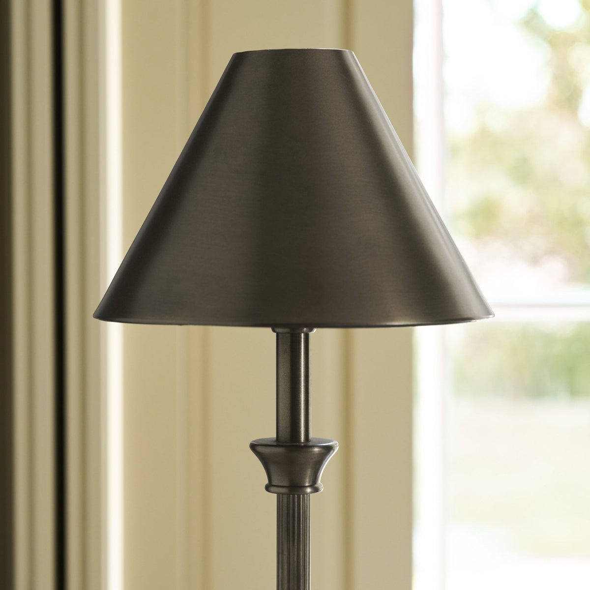 Close-up of a dark bronze lamp with a conical shade, against a blurred background of a window and light-colored wall.