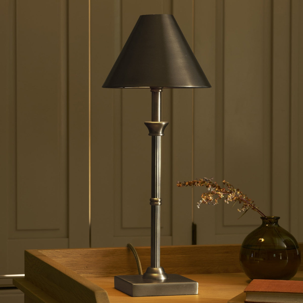 A tall, bronze table lamp with a dark shade sits on a wooden desk next to a brown vase with dried flowers.