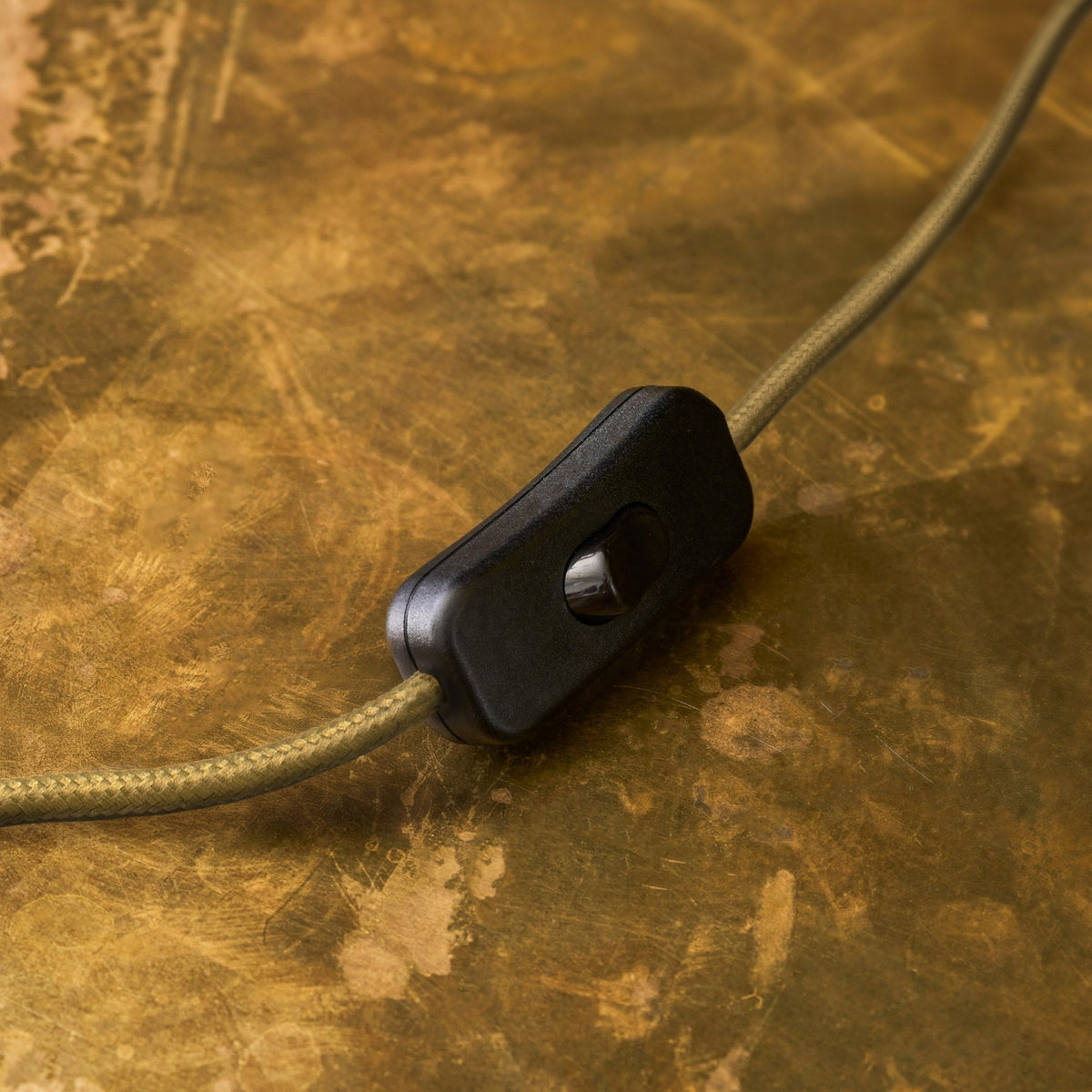 Close up of a black inline switch on a gold braided cable, set against a distressed brass background.