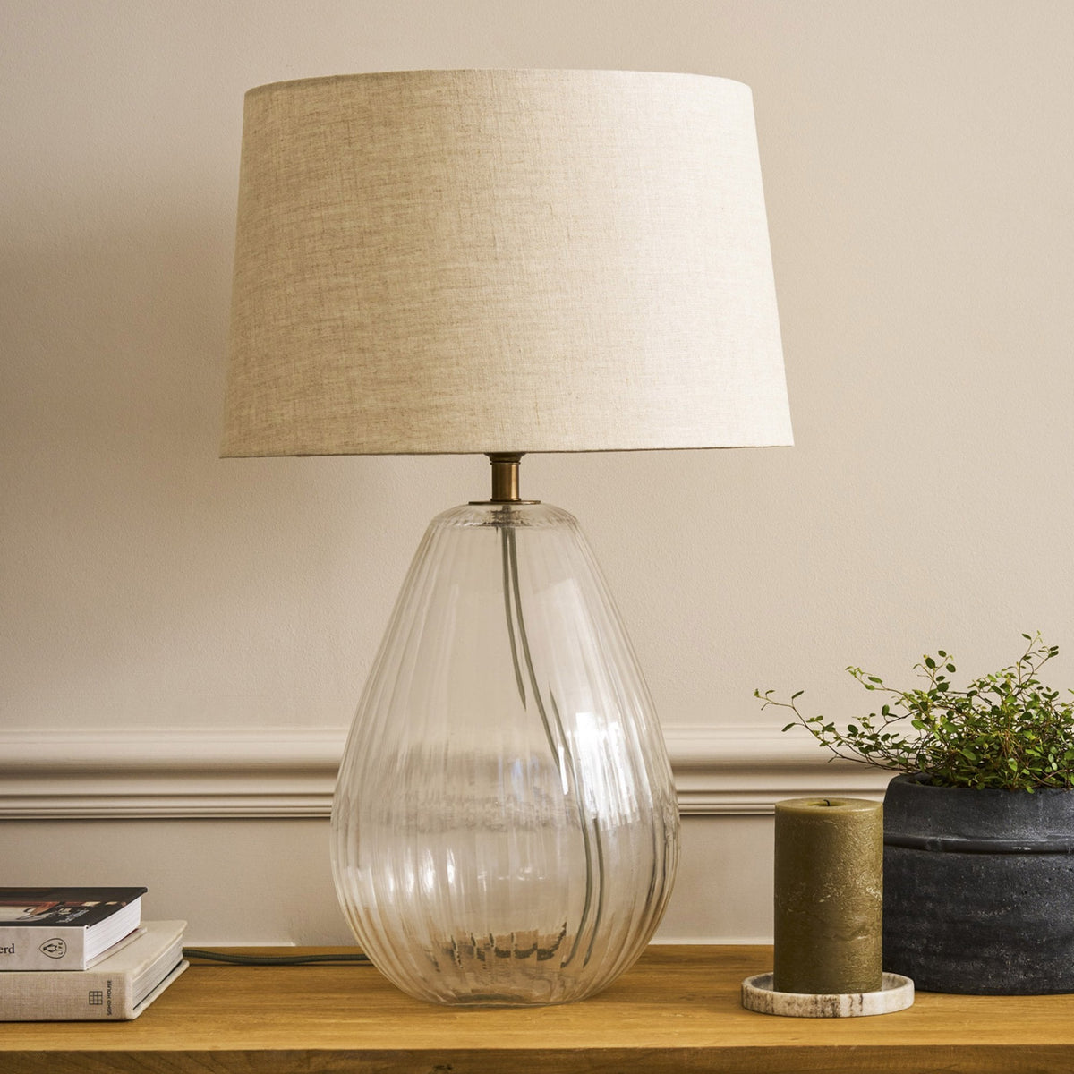 A clear glass table lamp with a ribbed base and a natural linen shade, on a wooden surface.