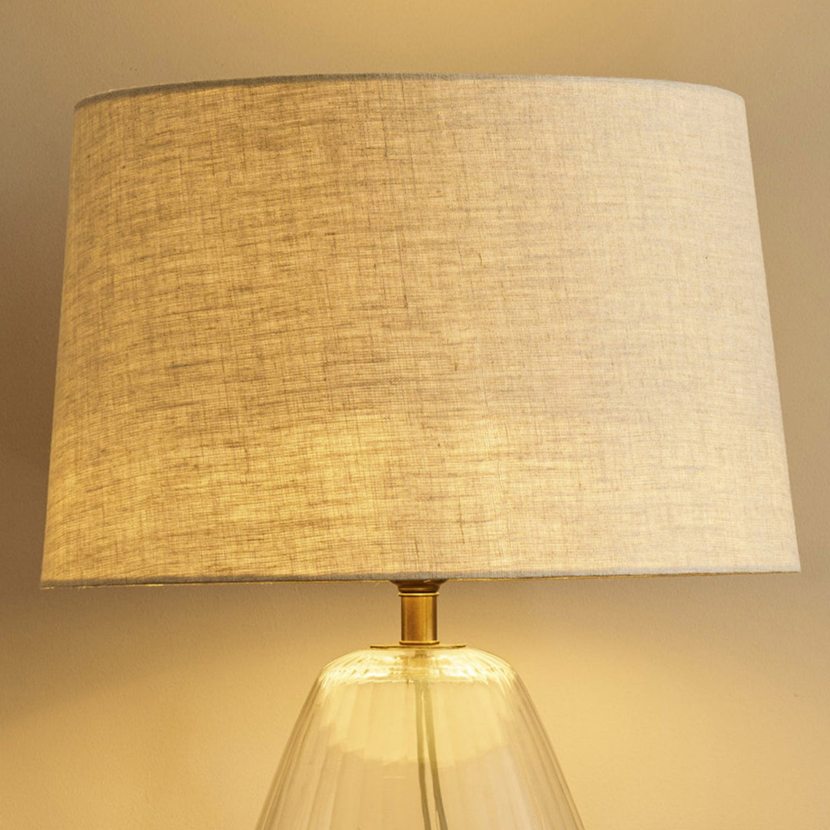 Close-up of a table lamp with a textured fabric shade and a clear glass base, illuminated with warm light. 