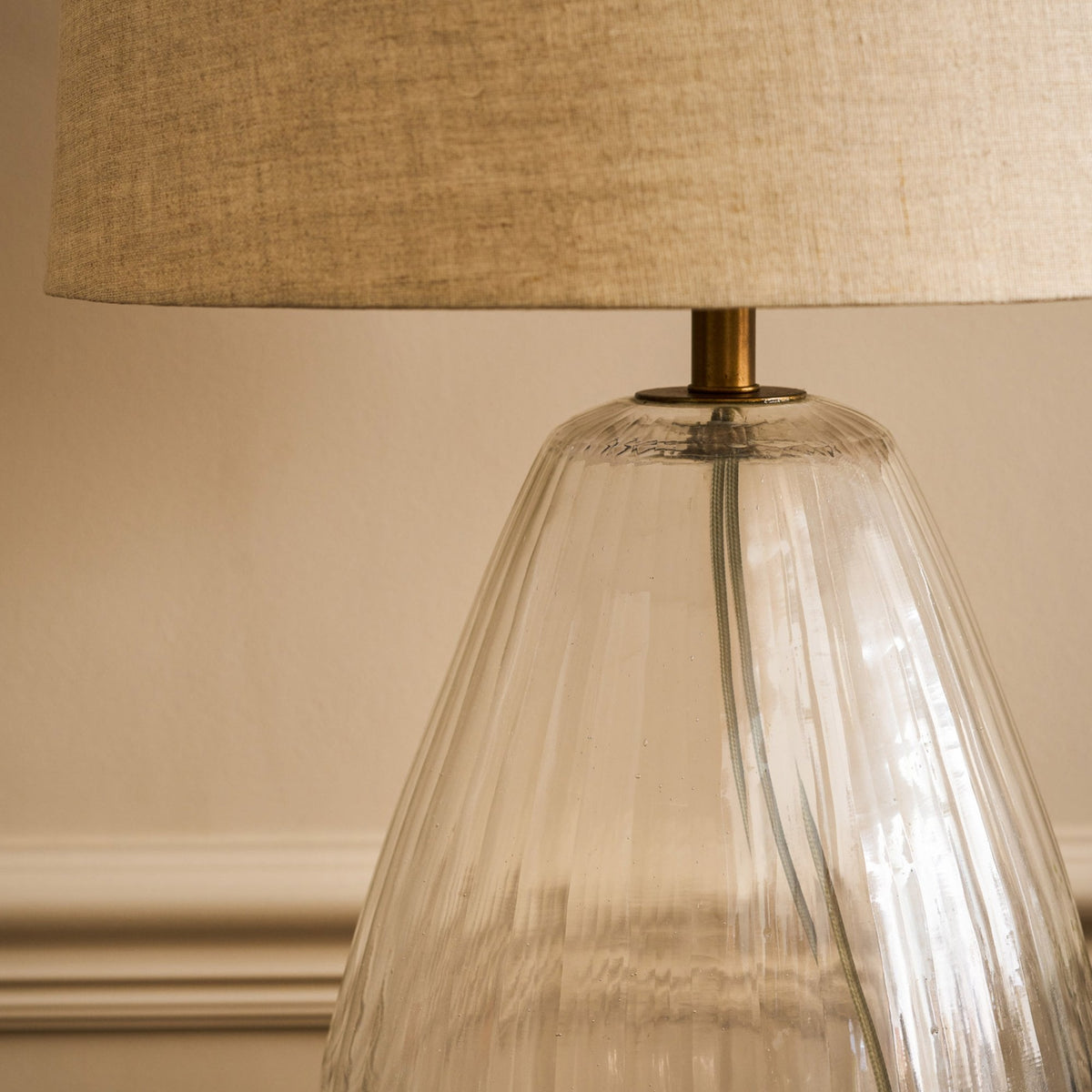 Close-up of a lamp with a clear, ribbed glass base and a light brown fabric shade. 