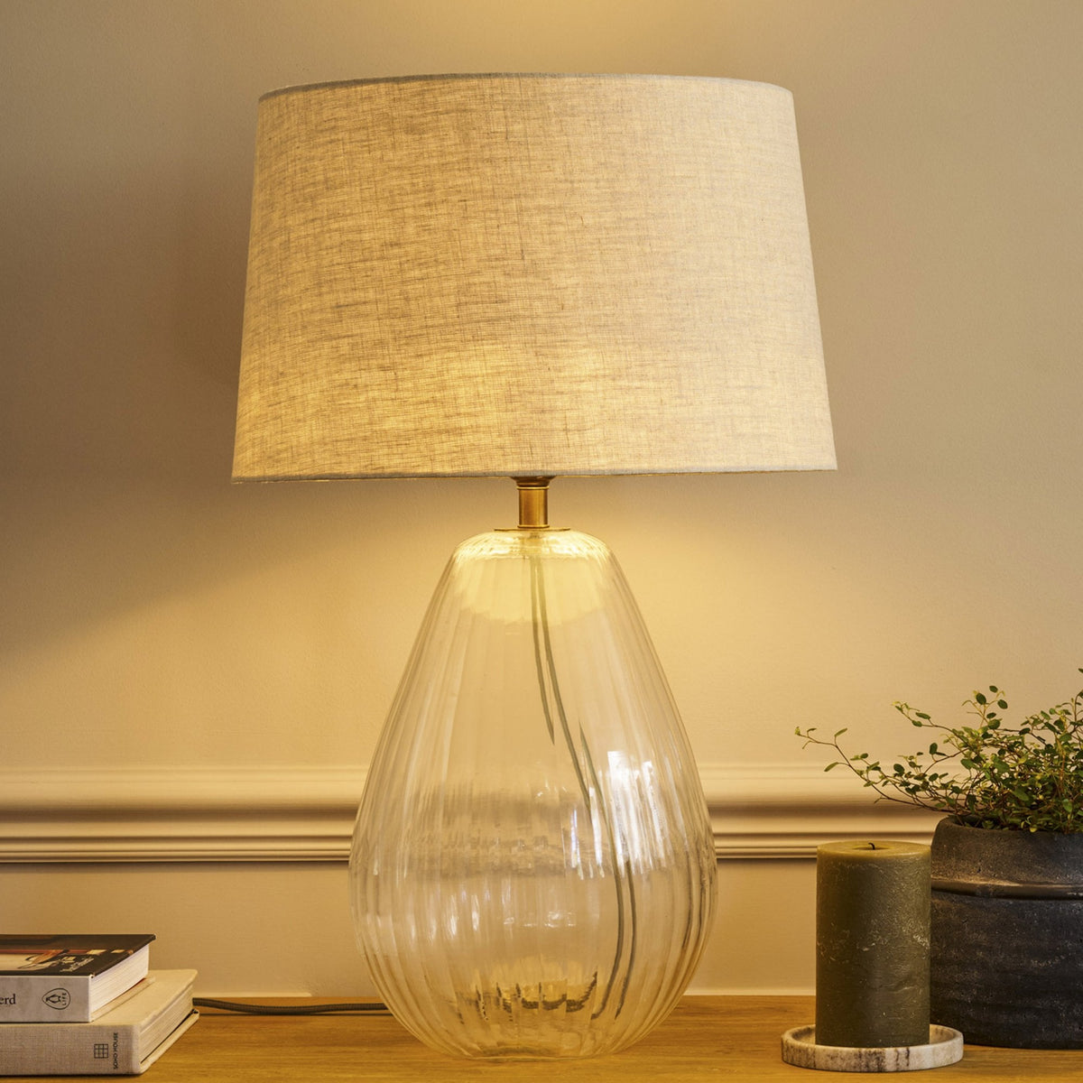 A clear glass table lamp with a ribbed base and a light grey fabric shade, illuminated on a wooden surface.