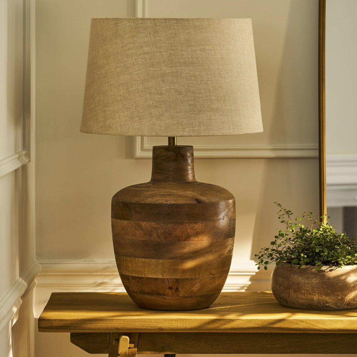 A wooden table lamp with a linen shade sits on a wooden console table next to a potted plant.