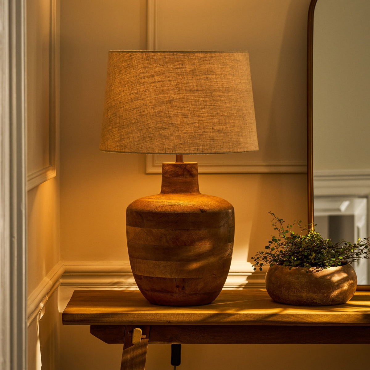 A wooden table lamp with a linen shade and a small potted plant on a wooden console table.