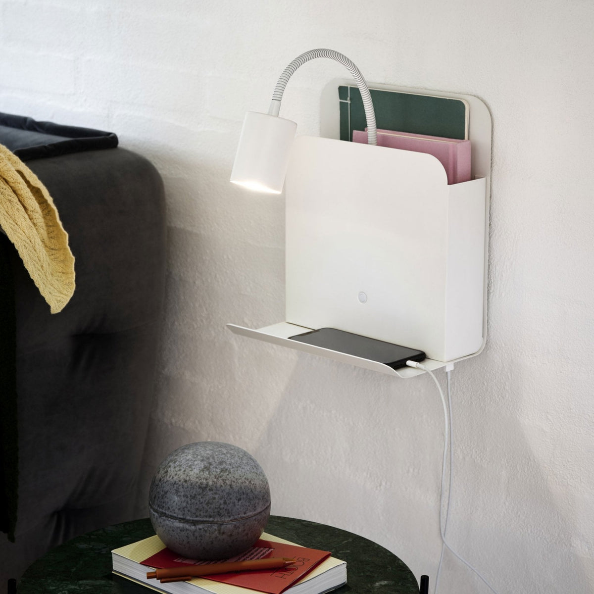 White wall shelf with light, phone, and books, next to a bed.