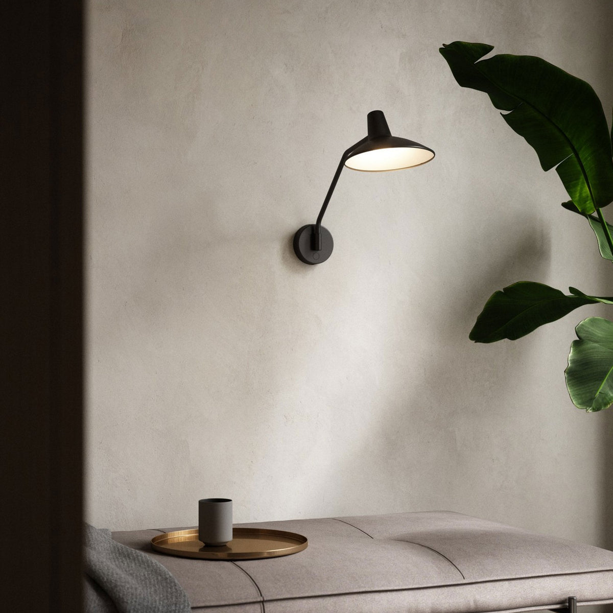Black wall lamp illuminates a beige wall above a bench with a tray, cup, and blanket