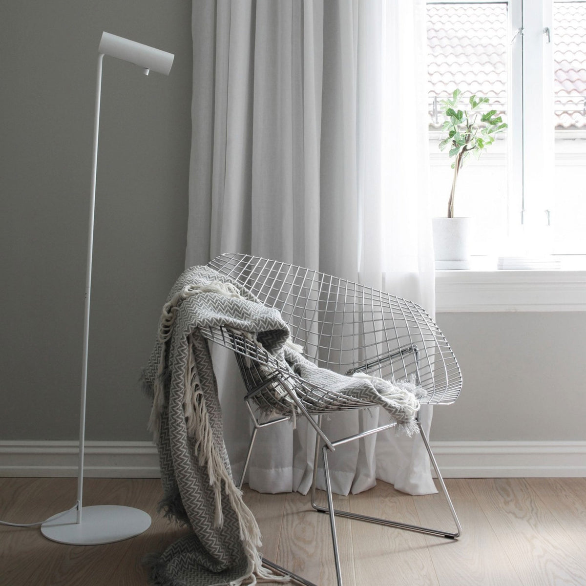 A white cylindrical floor lamp stands next to a white wire chair with a grey throw blanket.