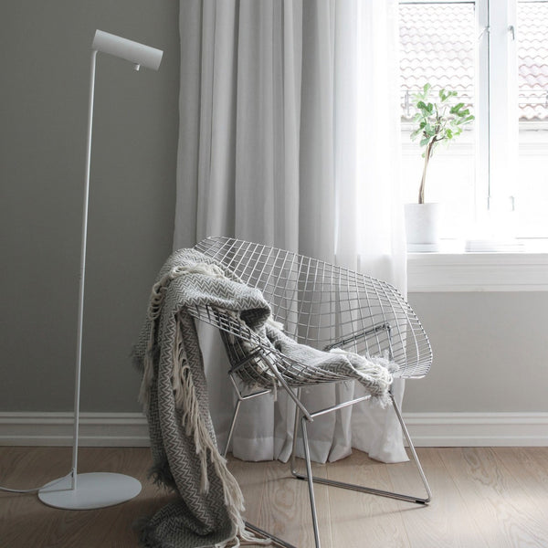 A white cylindrical floor lamp stands next to a white wire chair with a grey throw blanket.