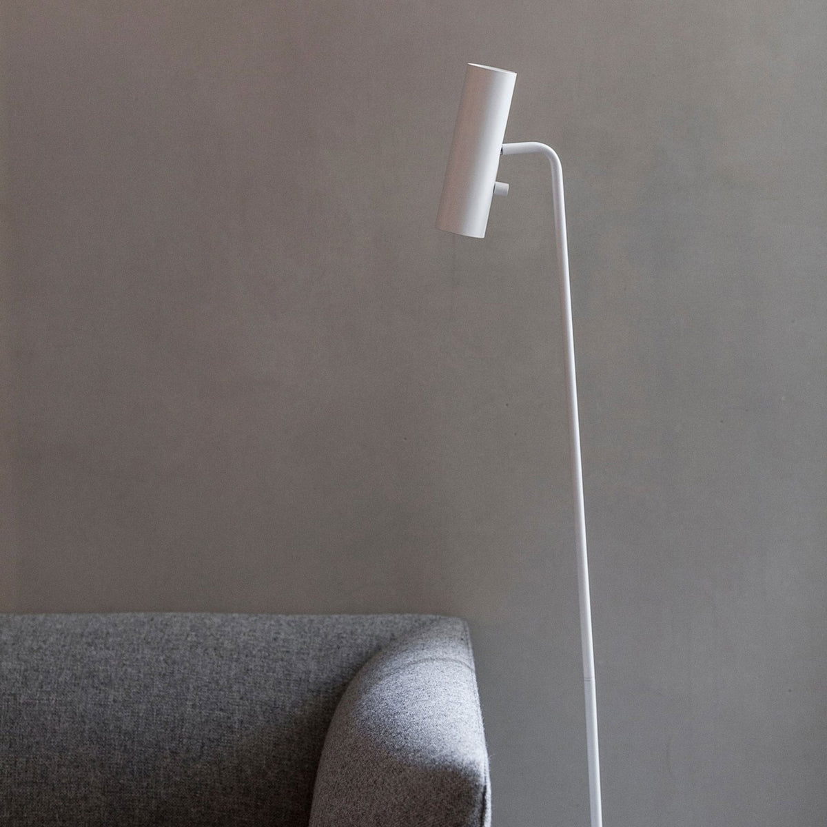 A white cylindrical floor lamp stands next to a grey sofa in a modern setting.