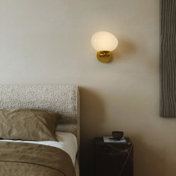 A white orb wall light with a brass base illuminates a bedroom setting.