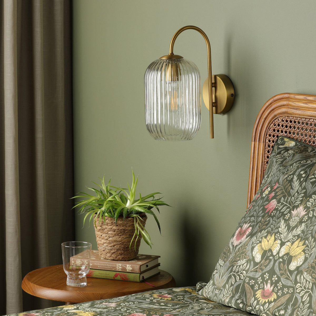 A brass wall sconce with a ribbed glass shade illuminates a bedroom with green walls and floral bedding.