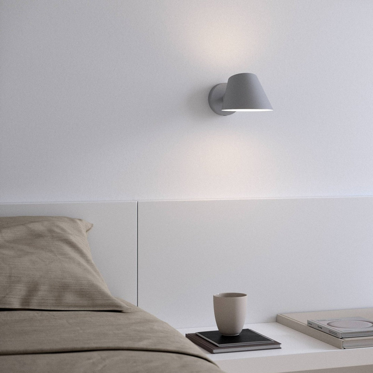 Grey wall lamp above a bedside table with a cup, book, and bed with brown pillows.