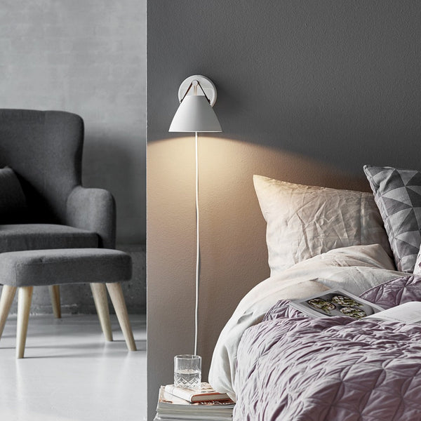 A white tapered wall light with a long white cord, illuminating a modern bedroom at night. 