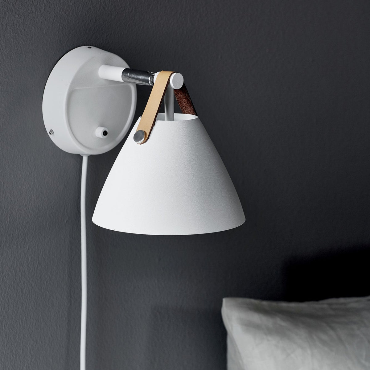 A white tapered wall light with a long white cord and a brown leather strap, mounted on a dark wall.