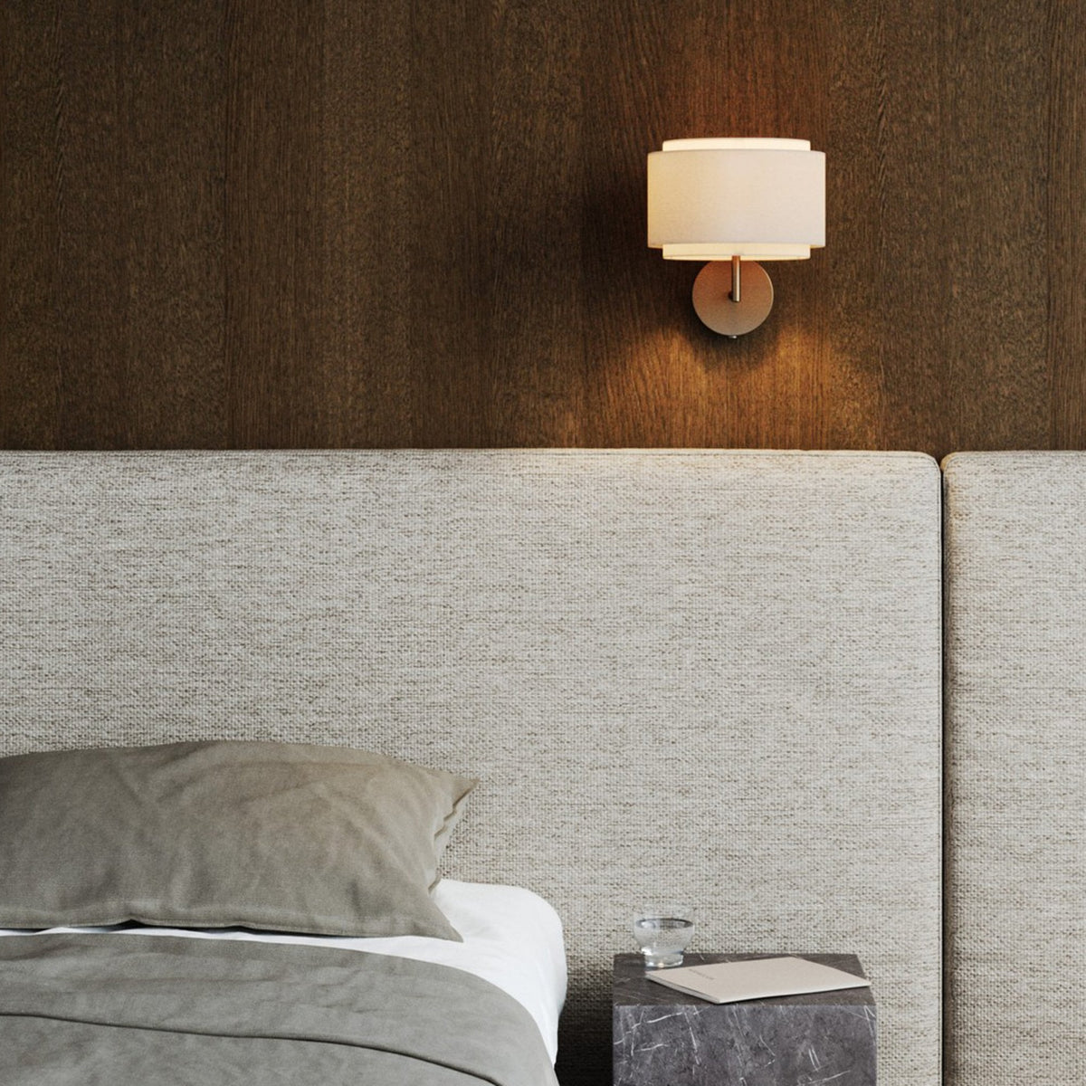 Hotel bedroom with an upholstered headboard and a wall light providing ambient lighting.