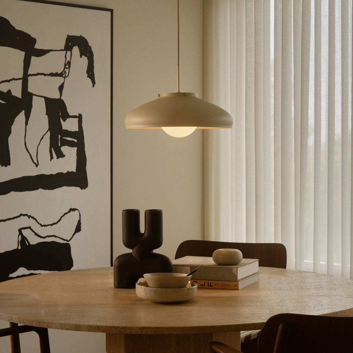 A beige pendant light hangs over a light wood table with bowls, books, and a dark vase