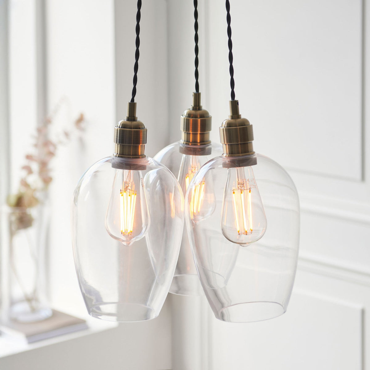 Three clear glass pendant lights with brass fittings hang against a window with natural light.